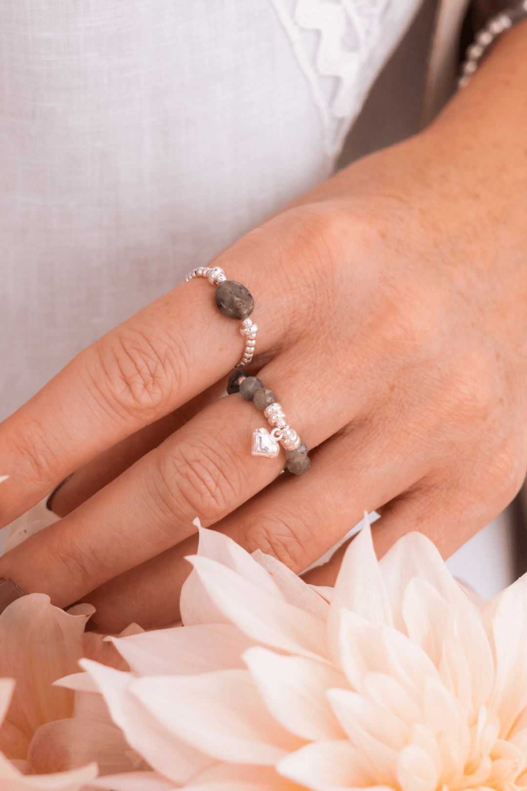 Bague élastique Be Tender en labradorite avec breloque cœur, portée sur un doigt, entourée de fleurs.