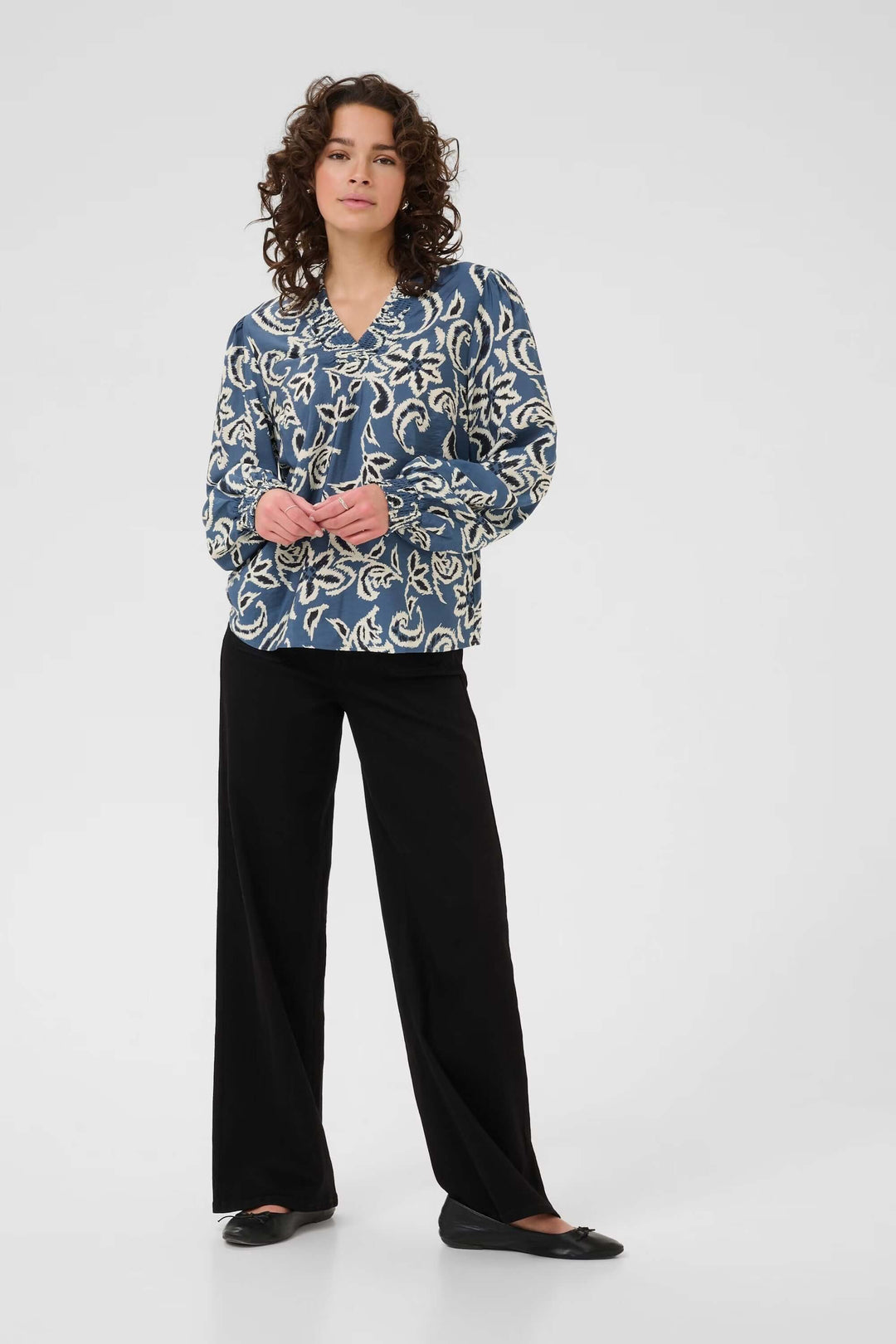 Woman wearing a blue and white floral print blouse with black pants, standing against a plain background.