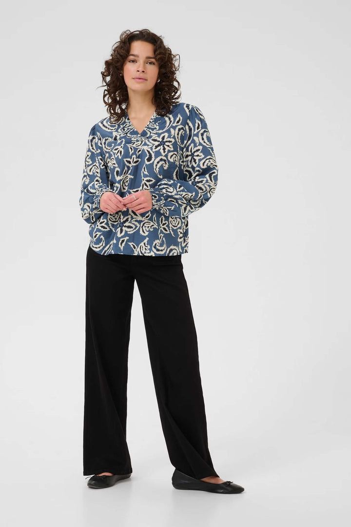 Woman wearing a blue and white floral print blouse with black pants, standing against a plain background.