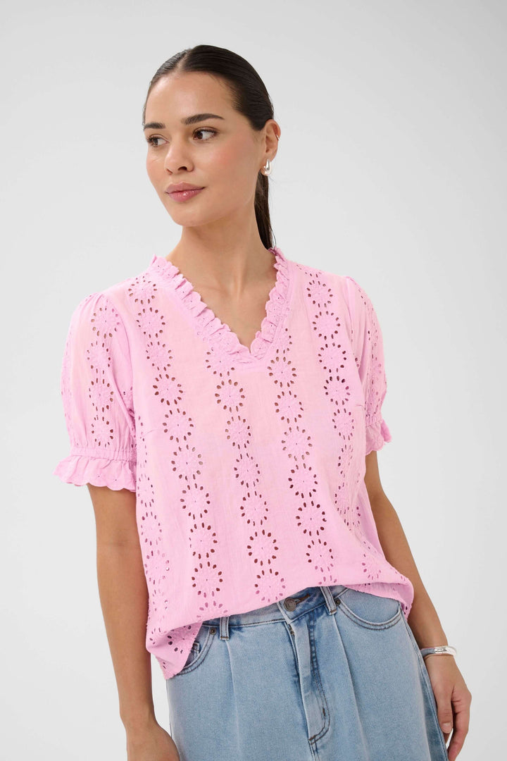 Blouse Dona brodée à manches courtes avec encolure volantée, broderies ajourées, portée avec un jean, look élégant et décontracté.