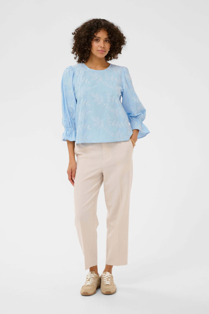 Femme portant la Blouse Marlee brodée à manches trois-quarts, en bleu avec broderies délicates, sur pantalon beige.