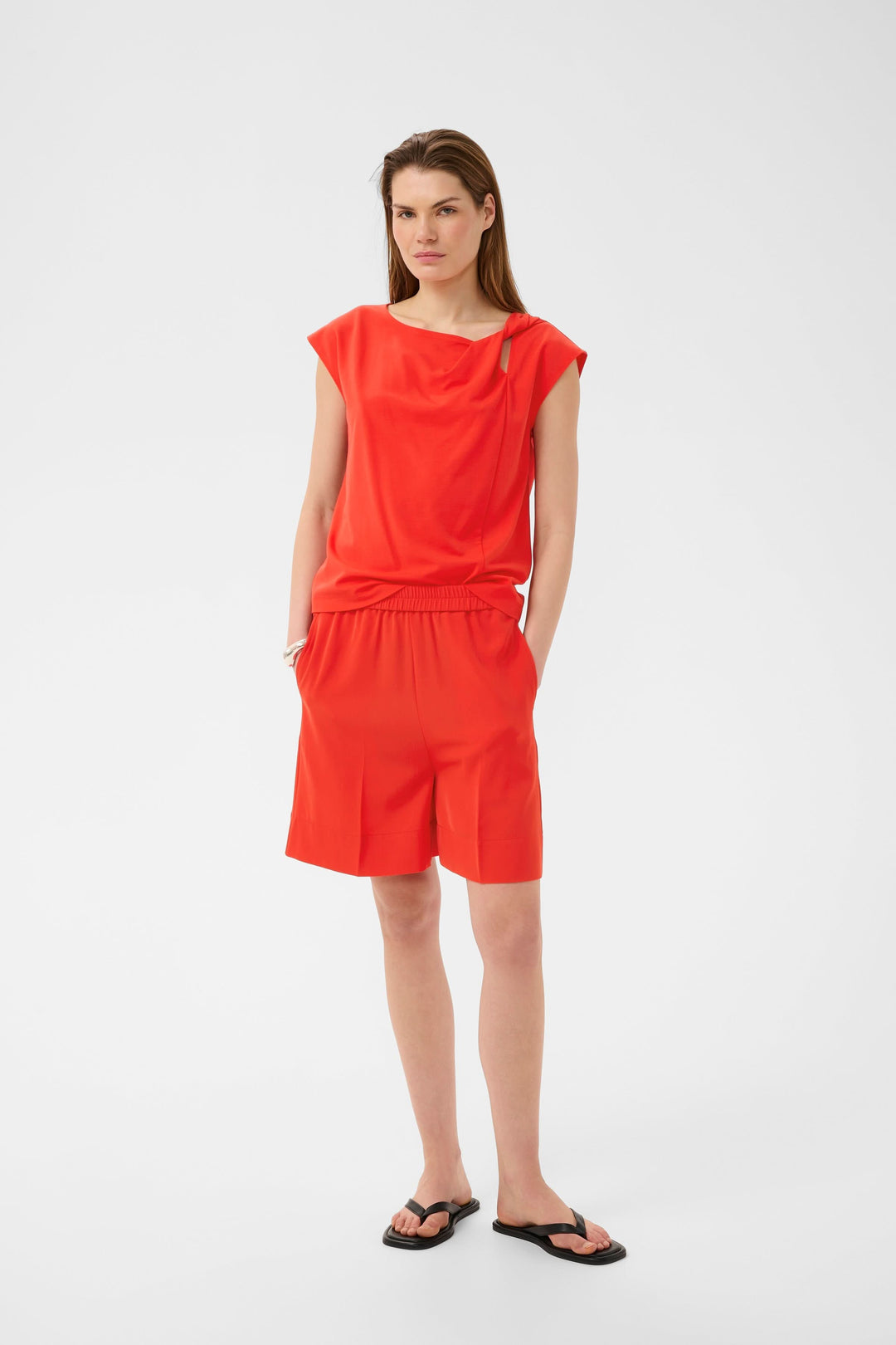 Blouse Pennie fluide à encolure drapée en rouge, tenue avec short assorti pour un style moderne et sophistiqué.