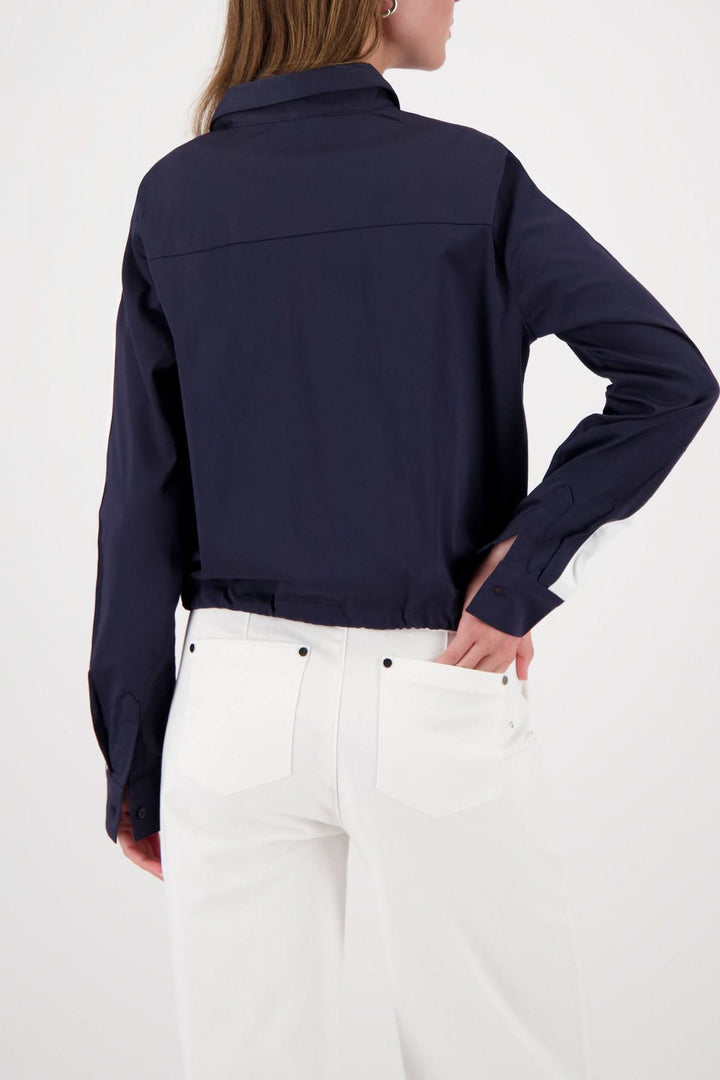 Blouse courte à cordon ajustable vue de dos, coupe moderne, col chemise, en bleu foncé, portée avec un pantalon blanc.