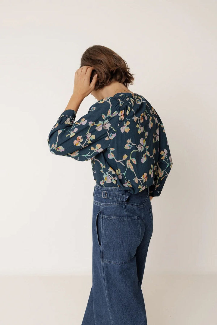 Femme portant une blouse florale à manches longues, dos à la caméra, style élégant avec jean, imprimé délicat et confortable.