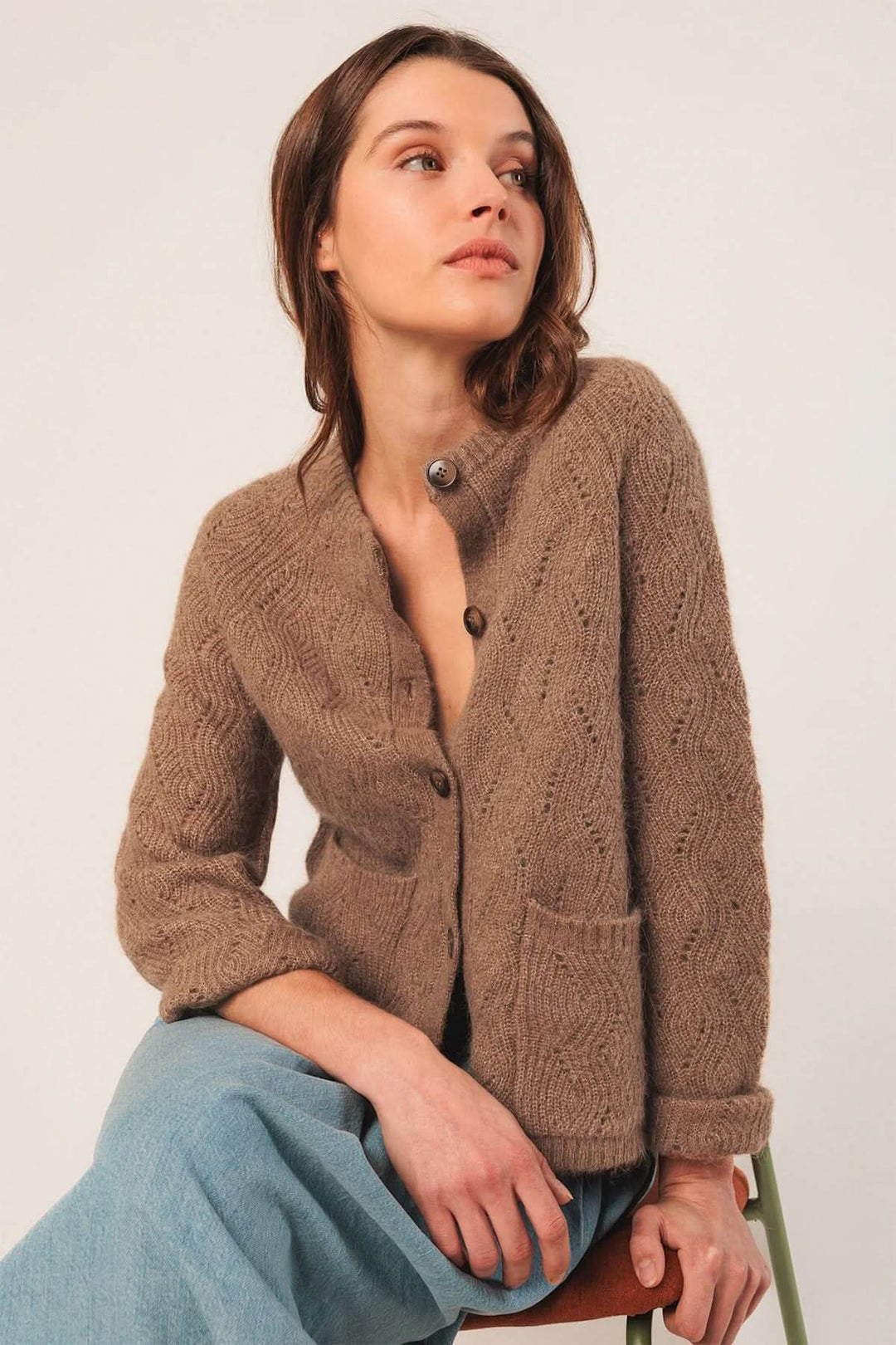 Femme portant un cardigan en maille texturée marron avec boutons et poches, assise sur une chaise.