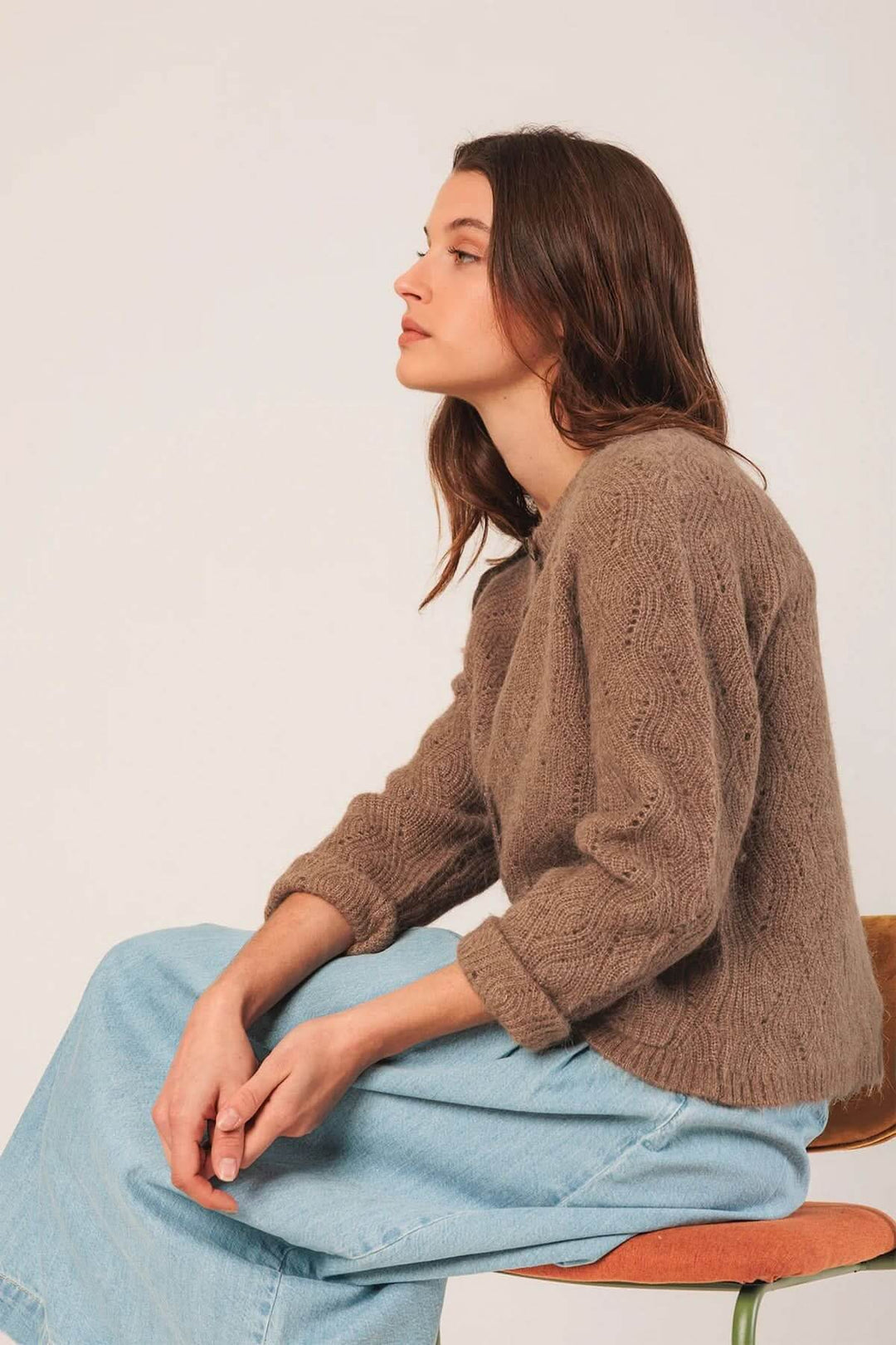 Femme portant un cardigan crochet en maille texturée, assise sur une chaise, avec un pantalon en denim.