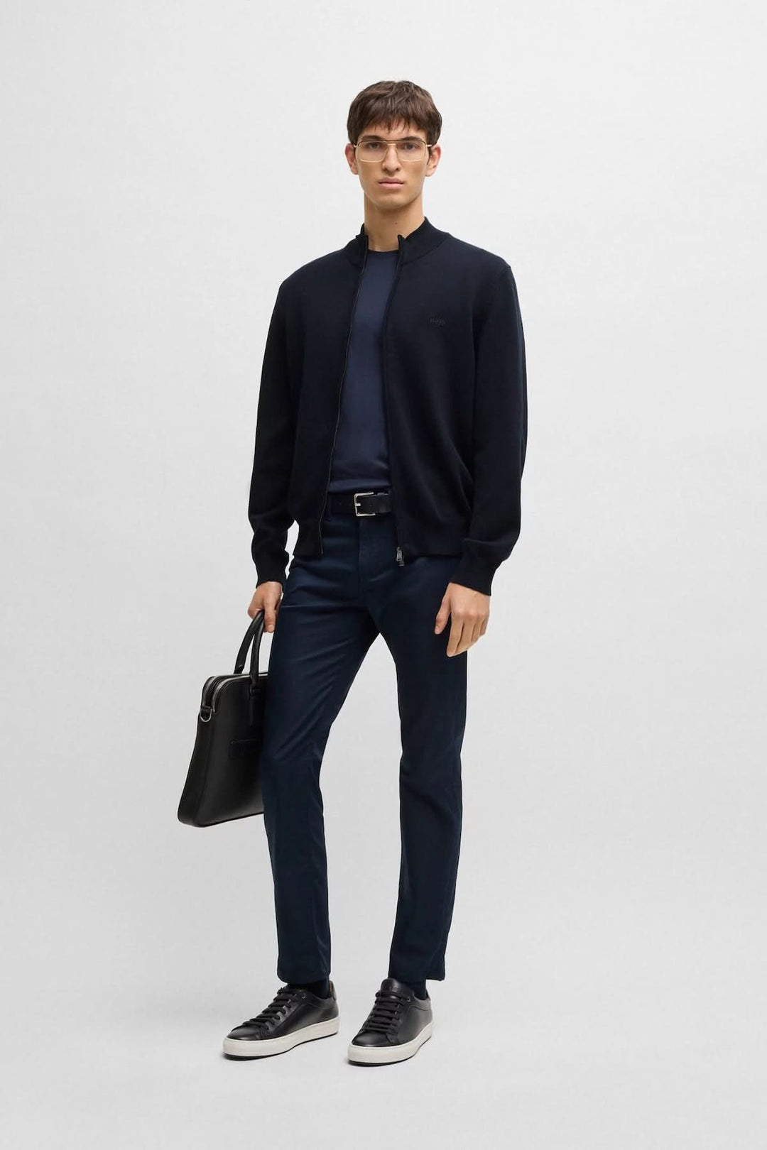 Homme portant un cardigan en maille noire avec col montant, jeans et sac, style décontracté et élégant.
