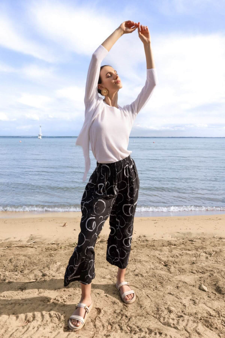 Femme portant un cardigan léger ouvert sur une plage, style décontracté avec pantalon noir à motifs, ciel ensoleillé en arrière-plan.