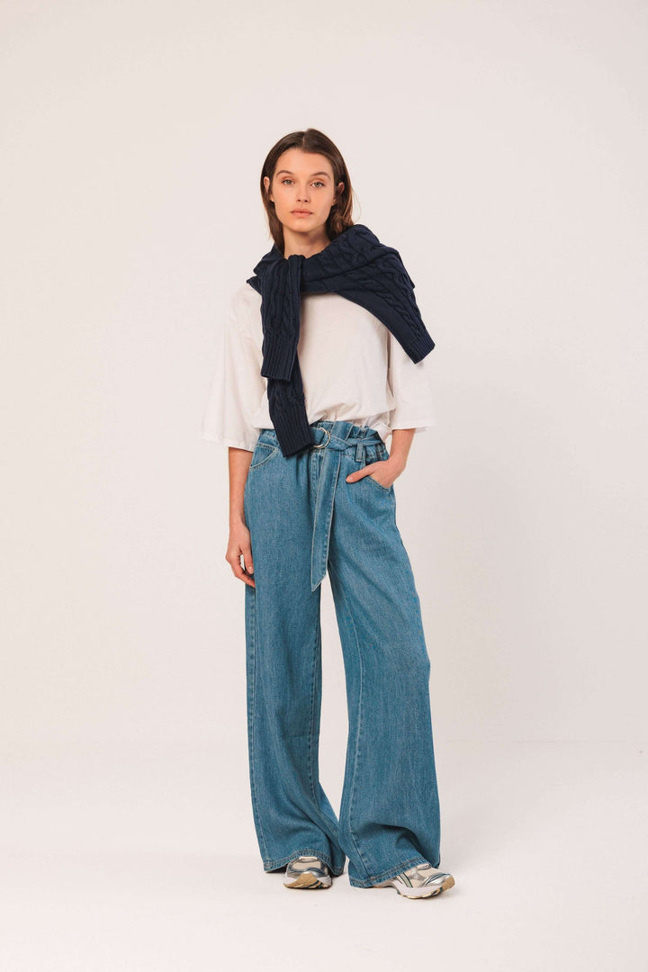 Femme en tenue décontractée avec un T-shirt blanc et un pantalon en denim large, portant un cardigan noué sur les épaules.
