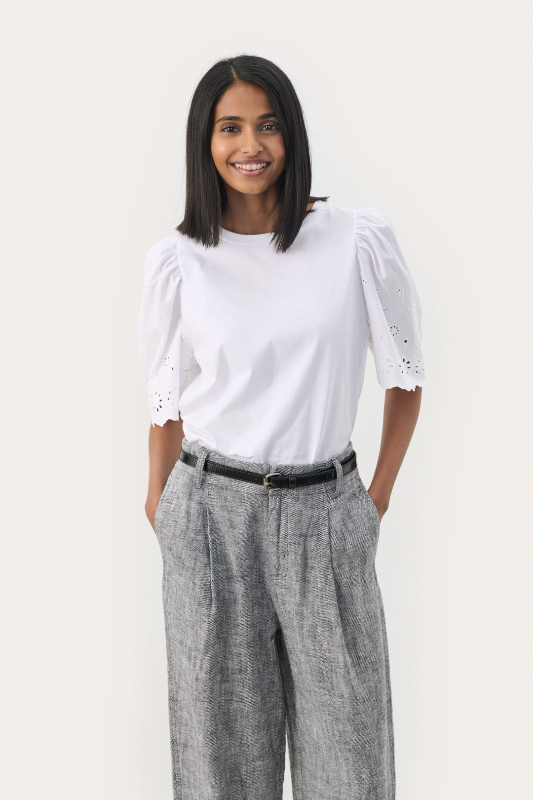 Femme souriante portant un t-shirt blanc avec des manches bouffantes et un pantalon gris, accessoirisée avec une ceinture.