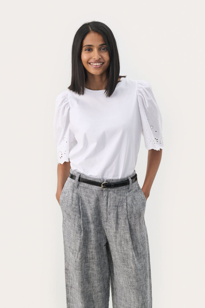 Femme souriante portant un t-shirt blanc avec des manches bouffantes et un pantalon gris, accessoirisée avec une ceinture.