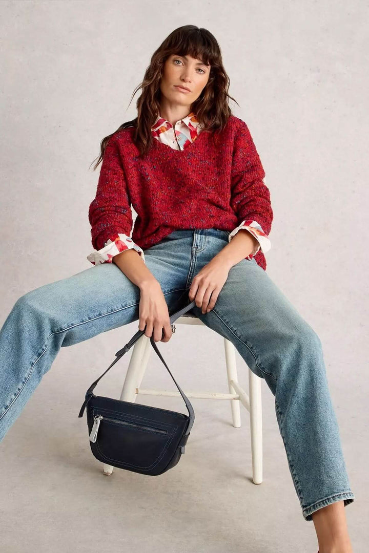 Modèle portant un chandail col en V rouge avec texture chinée, assis sur une chaise, jeans clairs et sac à bandoulière.