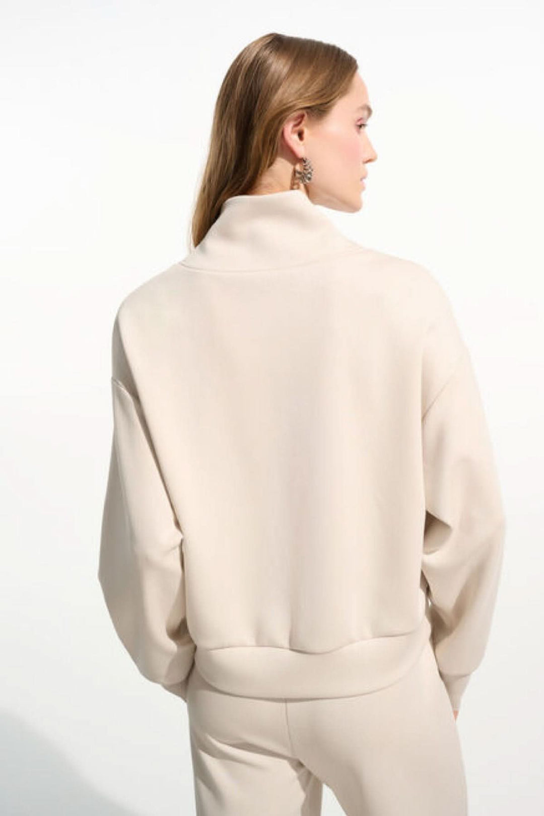 Femme portant un chandail à col montant beige avec fermeture demi-zip et coupe décontractée, vue de dos, style moderne et épuré.