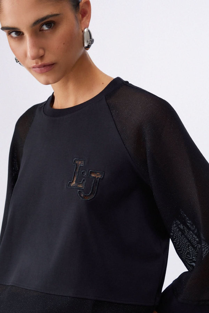 Chandail court à manches larges avec encolure ronde, coupe courte et manches trois-quarts, porté par un modèle féminin, écusson logo visible.