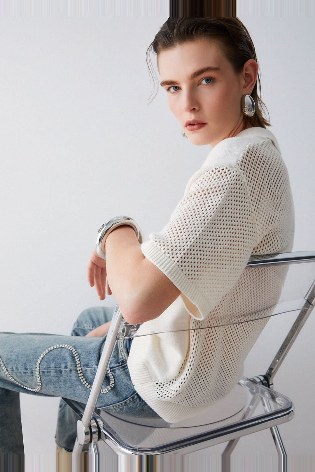 Personne portant un chandail en maille avec col polo et strass, assise sur une chaise, vêtement élégant et moderne.