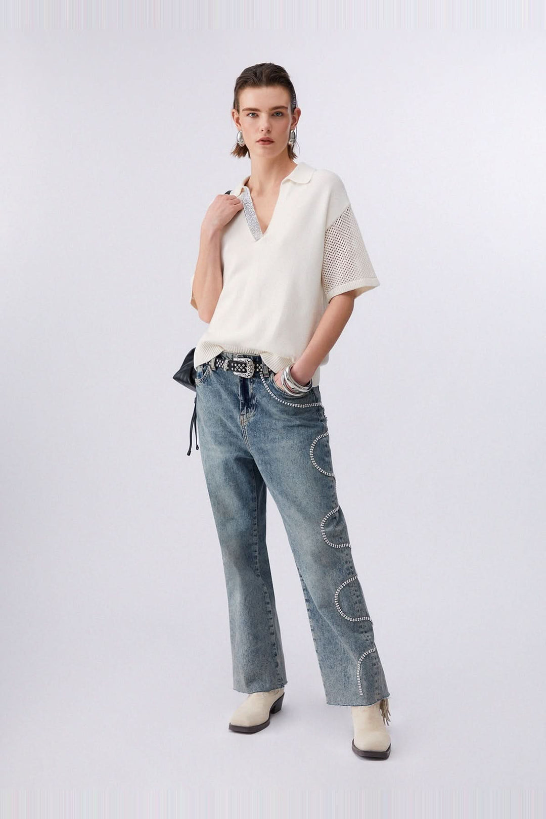 Chandail en maille avec col polo et strass, manches courtes et inserts ajourés, porté avec un jean et bottes blanches.