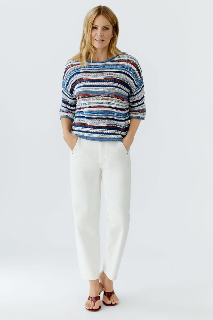 Femme portant un chandail en maille multicolore à manches trois-quarts avec un motif rayé, associé à un pantalon blanc pour un look décontracté.