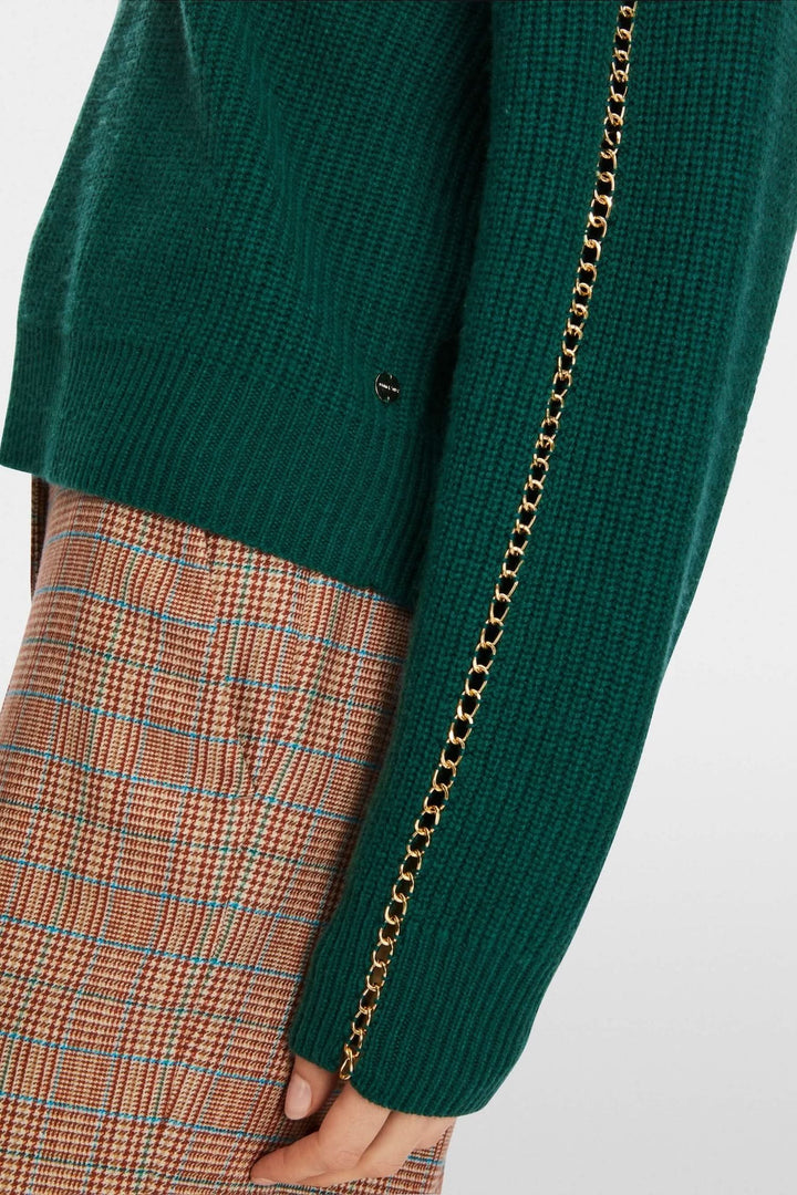 Pull en mailles vert avec chaîne métallique sur la manche, porté avec un pantalon à carreaux.
