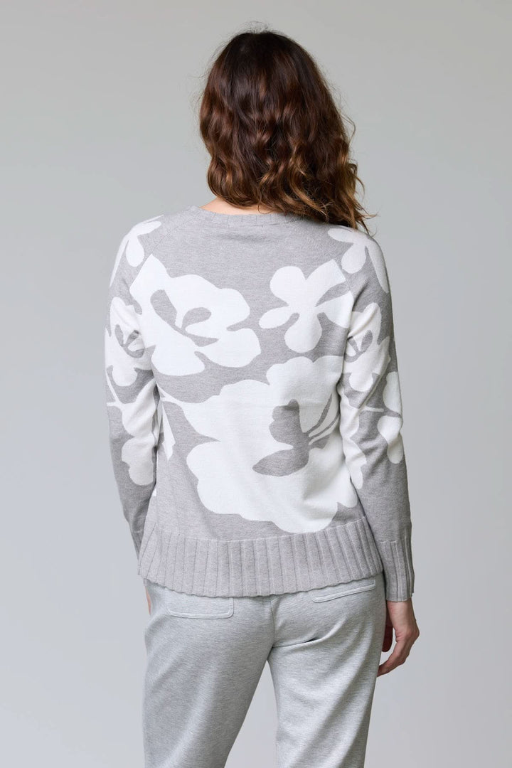 Femme portant un chandail en tricot à motif floral avec grandes fleurs blanches sur fond gris, vue de dos, coupe décontractée.