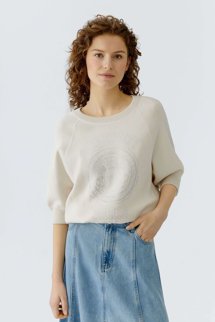 Femme portant un chandail à imprimé universitaire blanc avec manches trois-quarts, encolure ronde, sur jupe en jean.