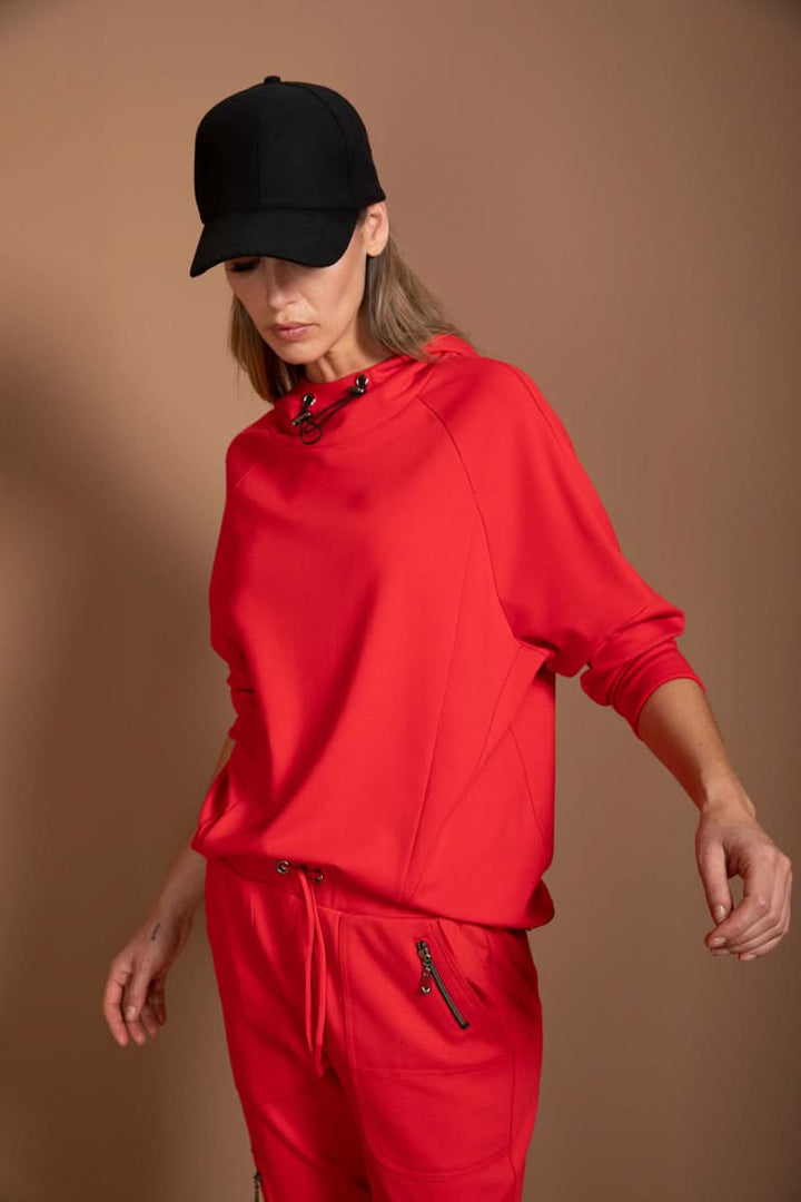 Chandail structuré rouge avec col montant et cordon ajustable, style athleisure, porté par une personne en casquette noire.