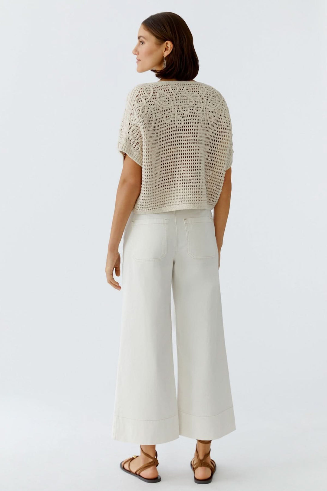 Femme portant un chandail tricot ajouré texturé beige avec un pantalon blanc, vue de dos, élégant et moderne, style décontracté chic.