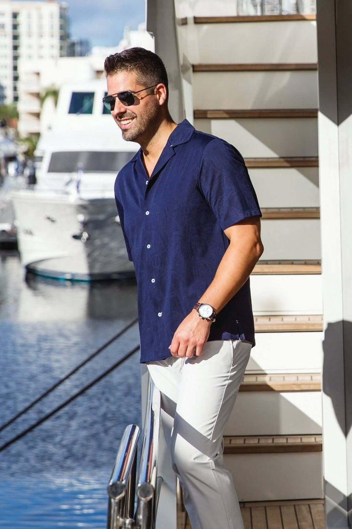 Homme portant une chemise à manches courtes bleu marine, lunettes de soleil, marchant au bord d'un quai.