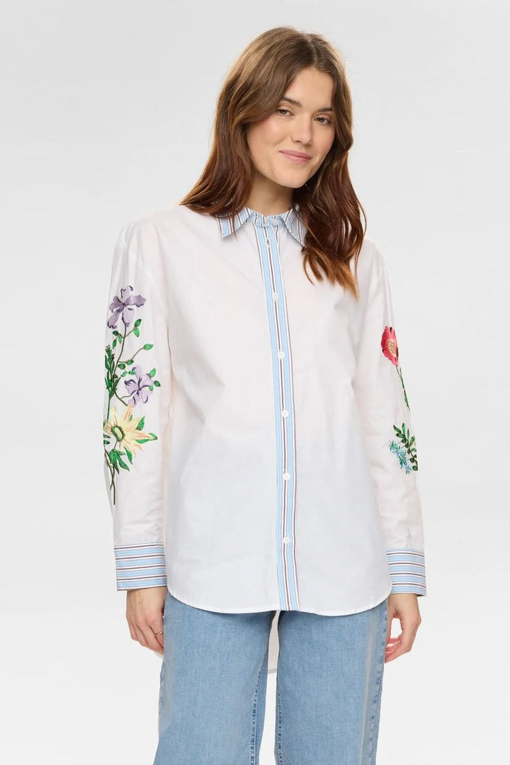 Chemise brodée à manches longues avec broderies florales sur les manches et col structuré, portée par une femme souriante.