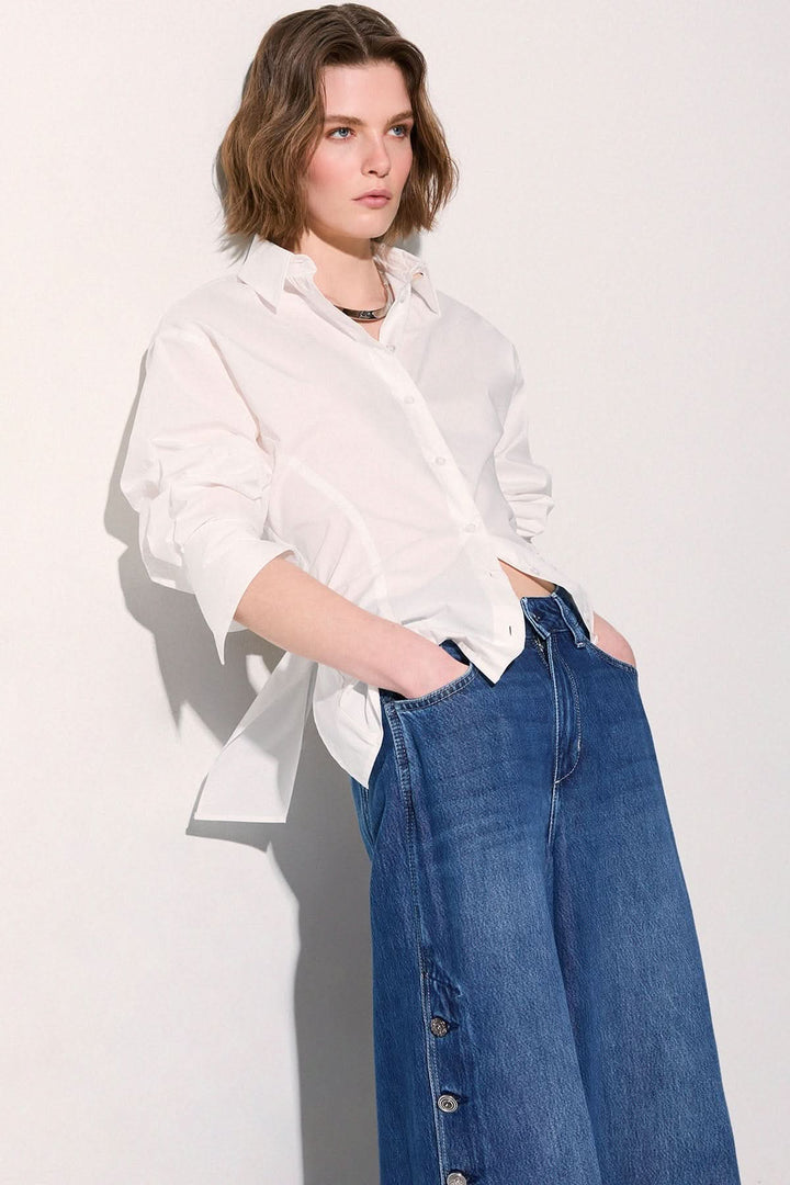 Femme portant une chemise à nouer au dos blanche, élégante et moderne, avec détails structurés, associée à un jean décontracté.