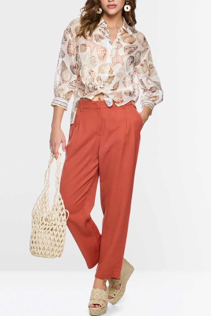 Chemisier fluide à imprimé coquillages avec pantalon rouille, style plage chic, femme portant un sac en osier et des sandales compensées.