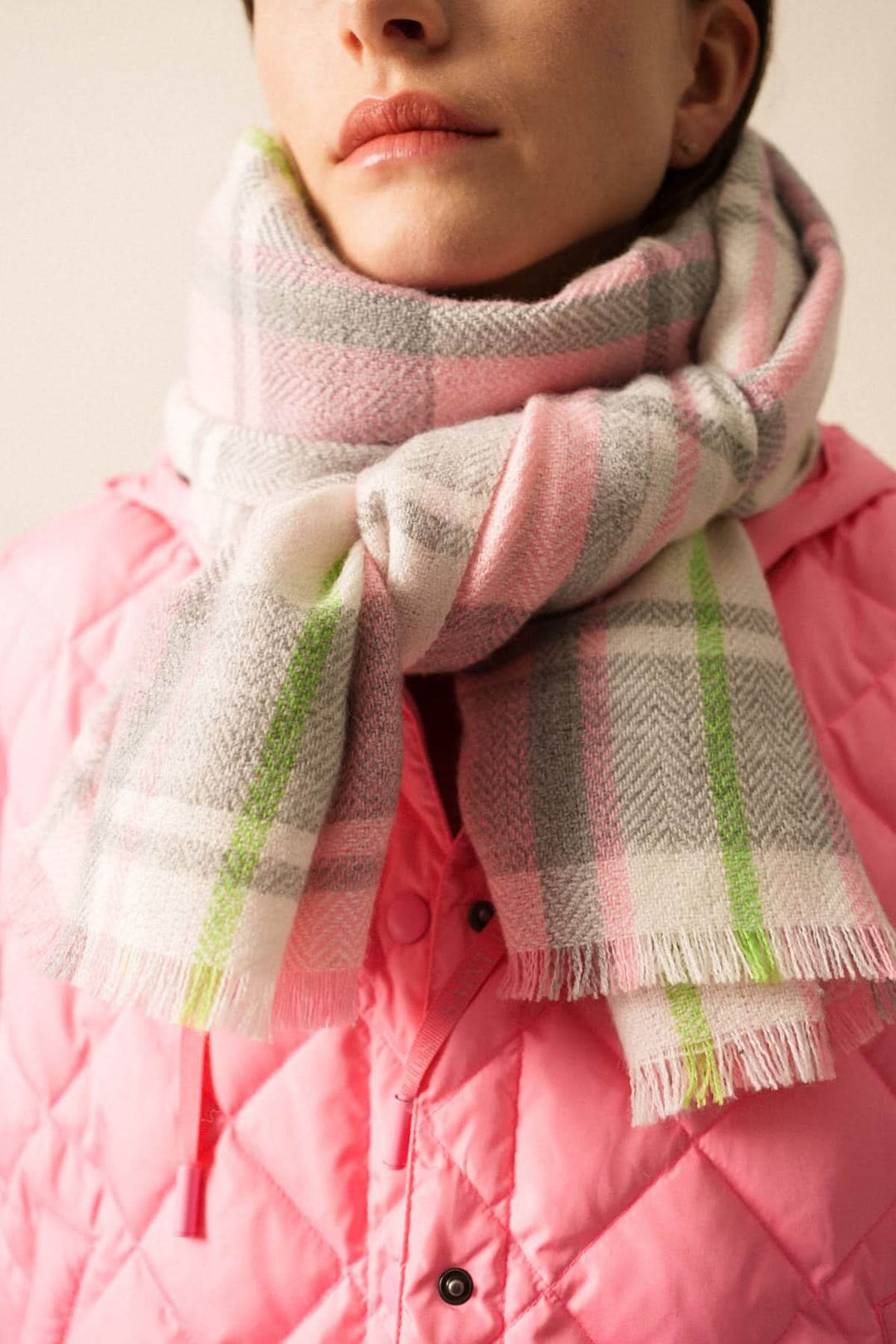 Foulard à carreaux doux et enveloppant porté par un modèle avec une veste rose, texture douce et chaude, détail frangé, motif classique.