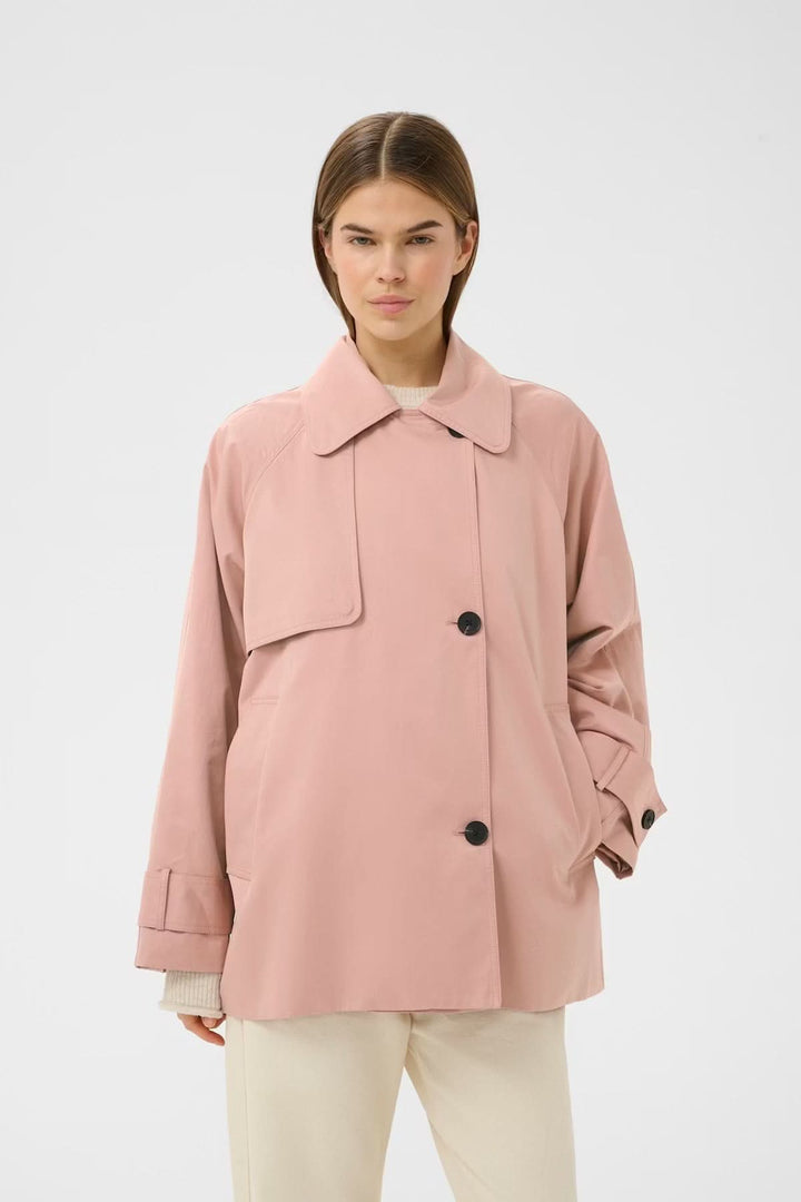 Femme portant un imperméable court à col large de couleur rose, coupe droite et boutons, idéale pour les tenues décontractées ou soignées.