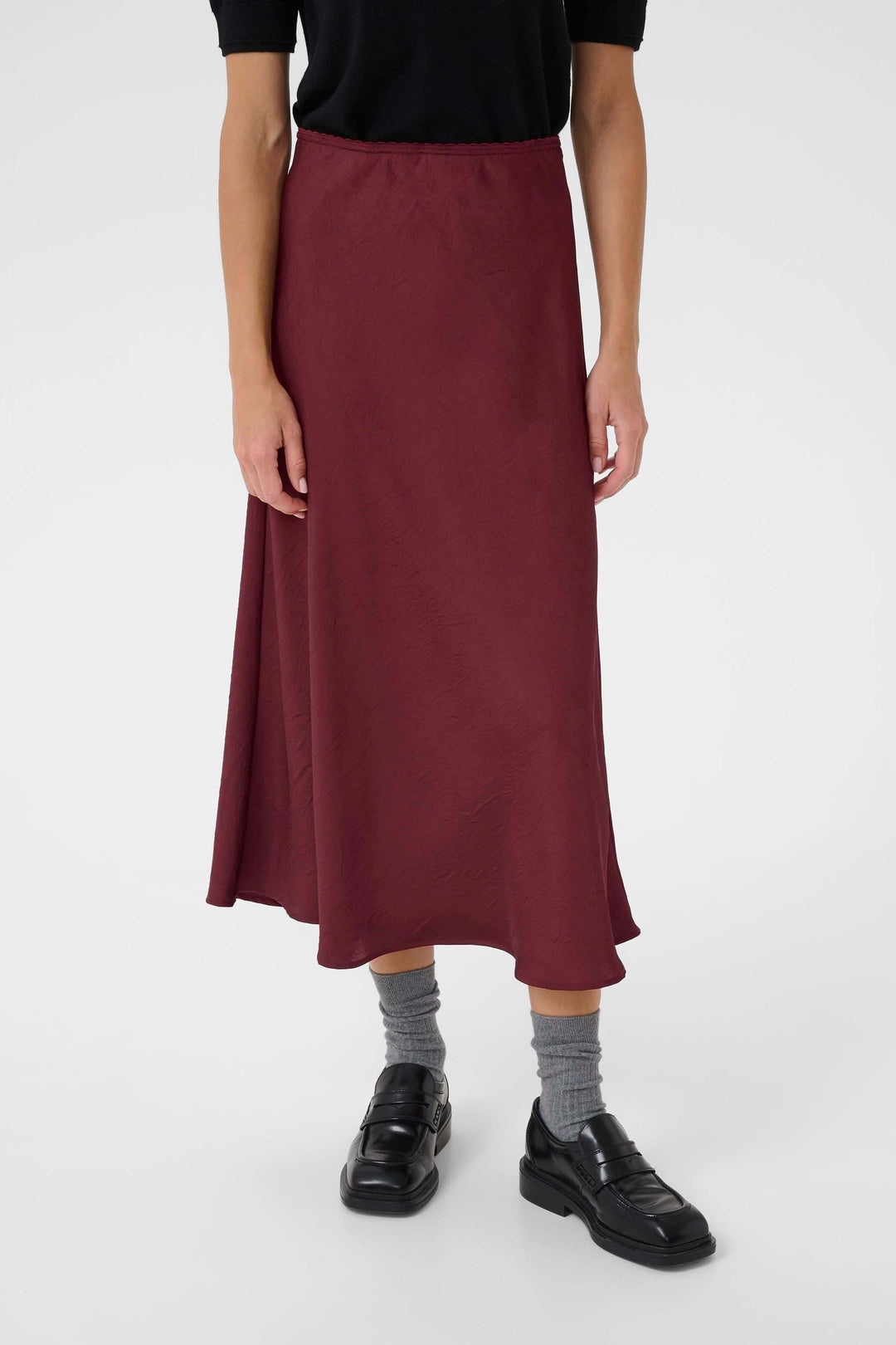 Jupe midi fluide rouge bordeaux avec coupe évasée, portée avec un haut noir et des chaussures noires.