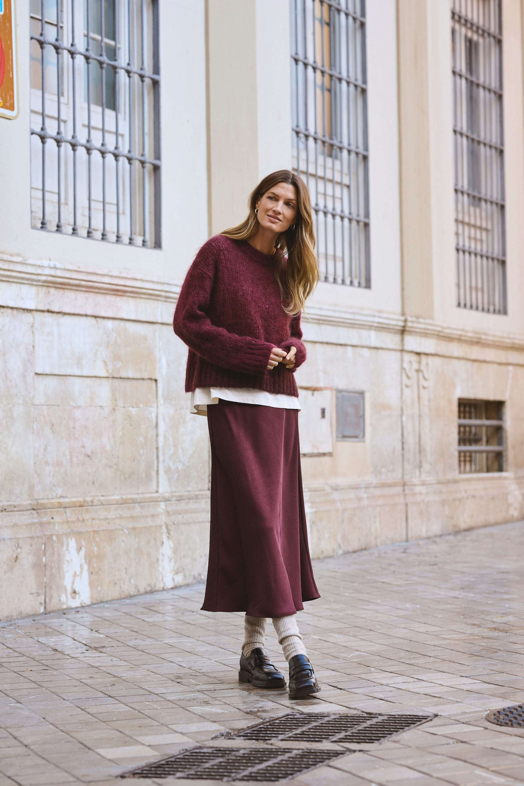 Femme portant une jupe midi fluide bordeaux et un pull oversize dans une rue élégante.