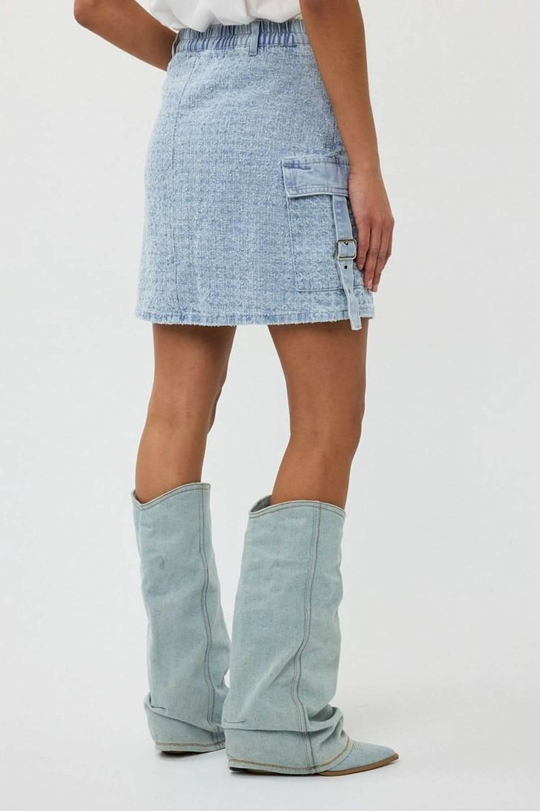 Jupe en denim texturé avec poche cargo latérale, coupe trapèze et sangle ajustable, portée avec des bottes.