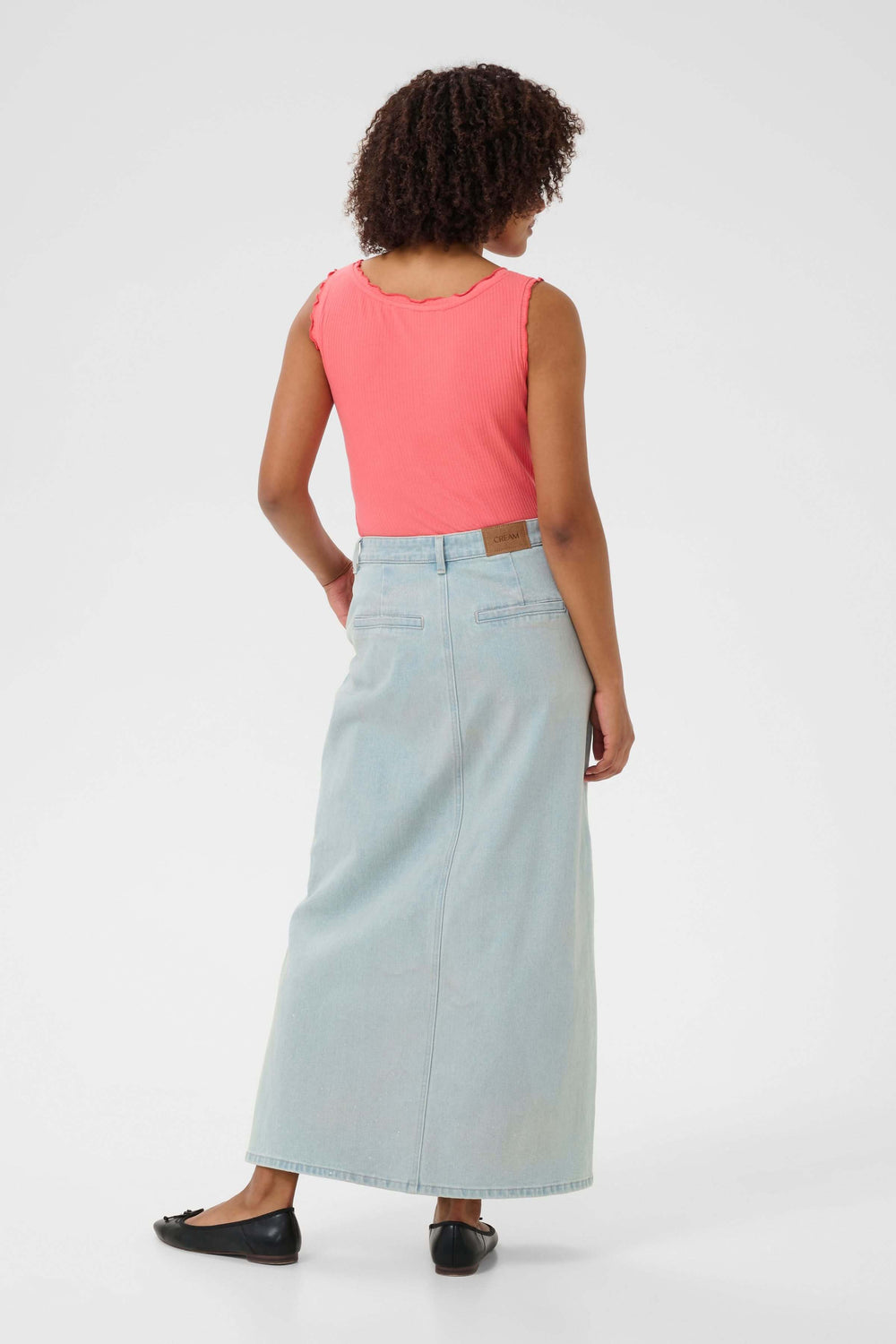 Femme vêtue d'une jupe longue en denim clair, vue de dos, avec un haut rose et des chaussures noires.