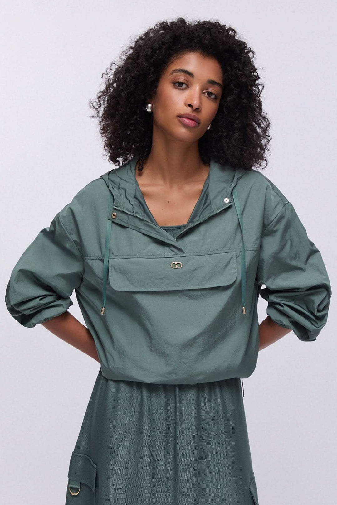 Femme portant un manteau coupe-vent à capuche avec poche poitrine, style moderne et décontracté, couleur vert forestier.
