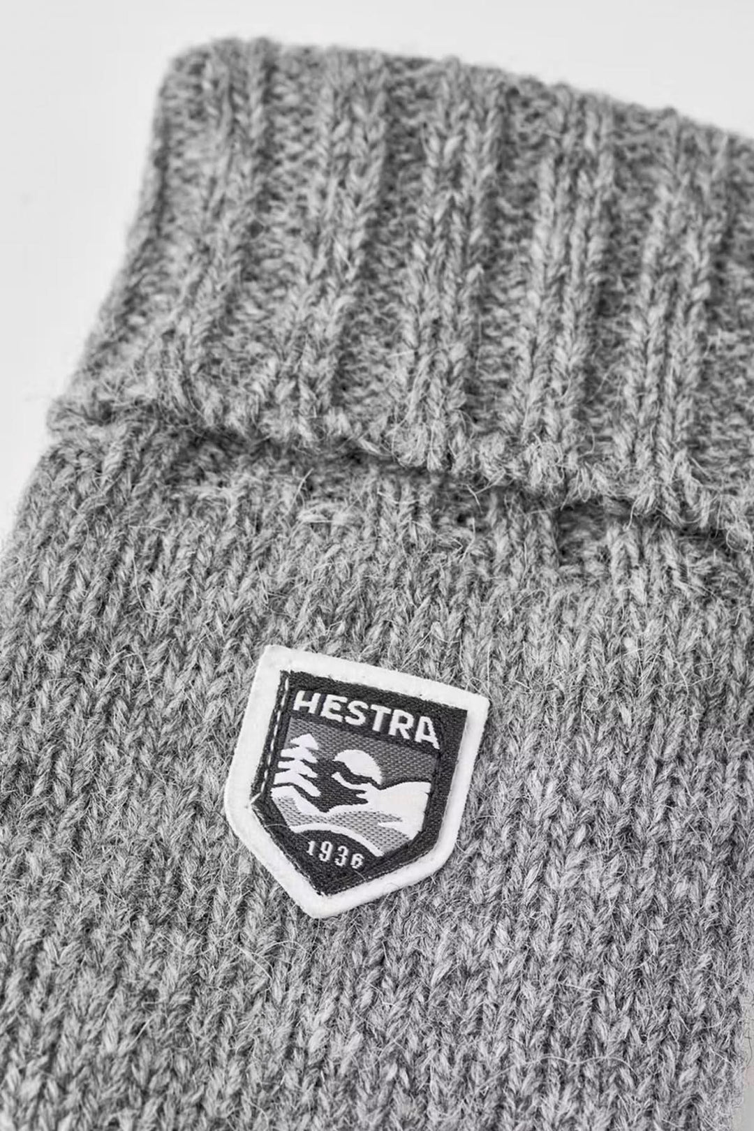 Mitaine en laine basique avec logo Hestra sur tissu gris tricoté, offrant chaleur et confort.