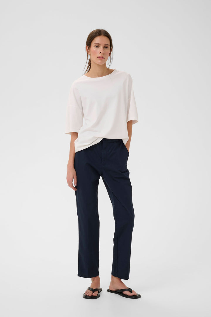 Femme portant un pantalon Diaz droit épuré bleu marine, coupe moderne et décontractée, associé à un haut blanc et des sandales noires.