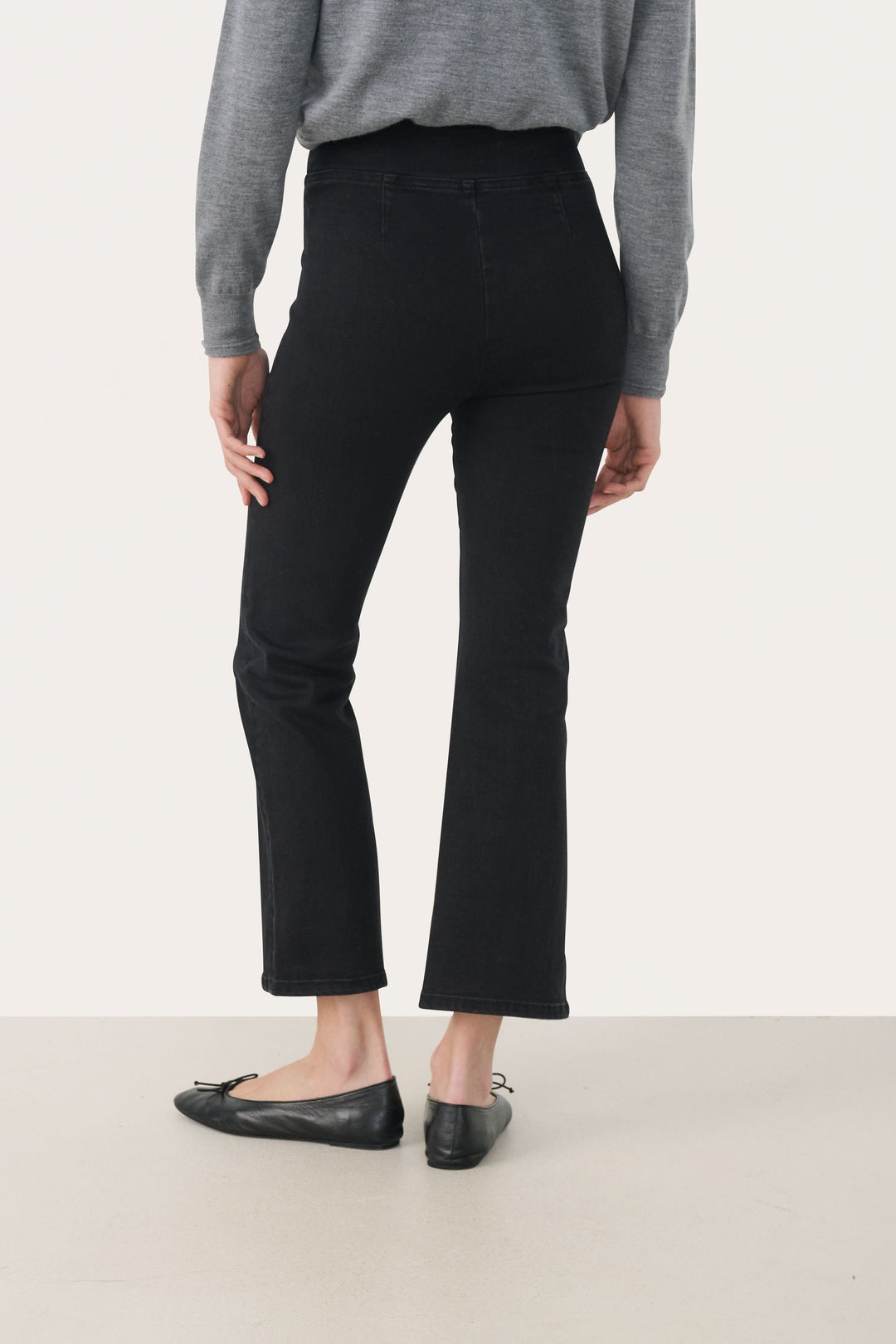 Femme portant le Pantalon Nikela noir évasé à taille haute avec un pull gris et des ballerines noires, vue de dos.