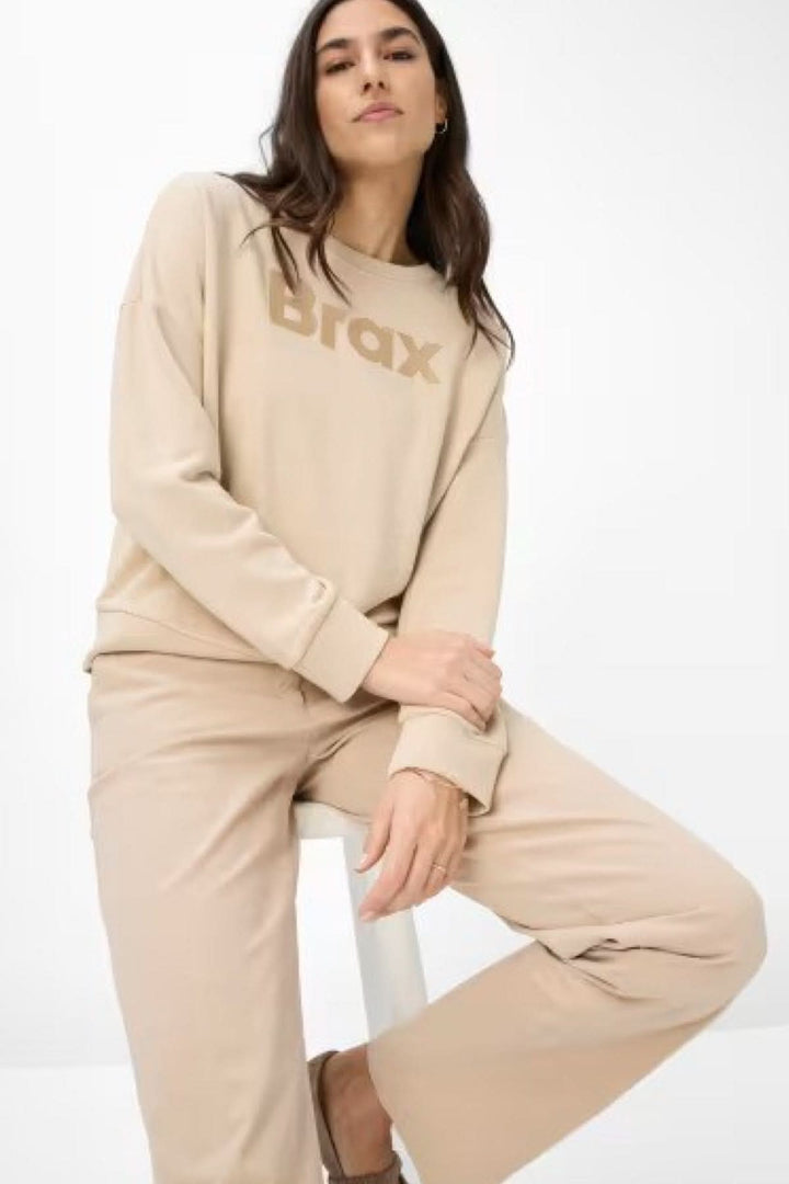 Femme portant un pull beige Brax et un pantalon chino assorti, posant de manière décontractée.