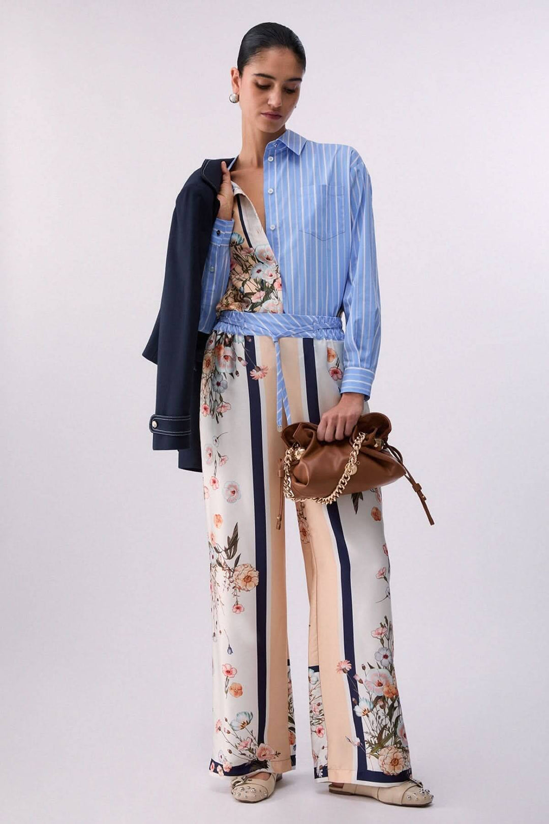 Femme portant un pantalon fluide à motifs et rayures contrastées avec une chemise rayée bleue, tenant un sac marron mode.