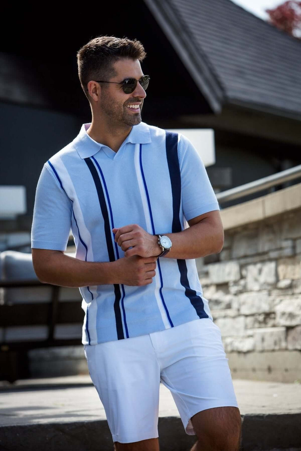 Homme portant un polo à rayures bleues et blanches, avec des shorts blancs, souriant au soleil.