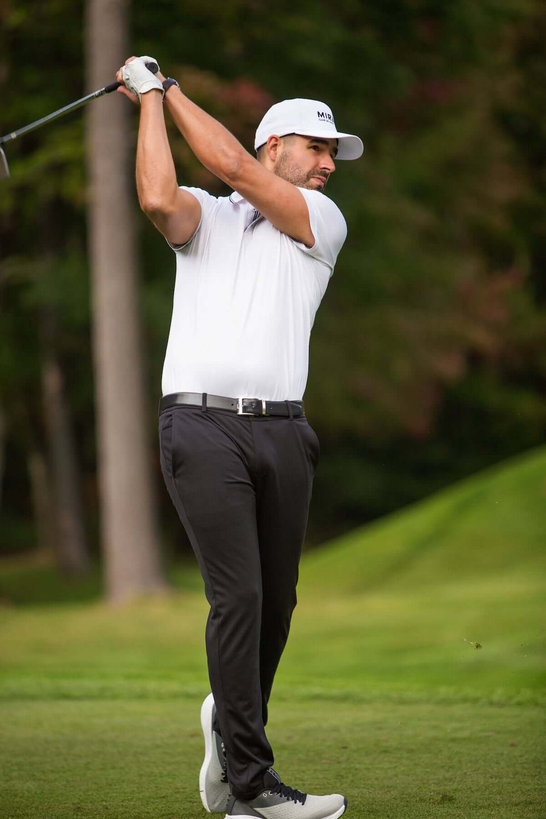 Homme jouant au golf, portant un polo blanc et un pantalon noir sur un terrain verdoyant.