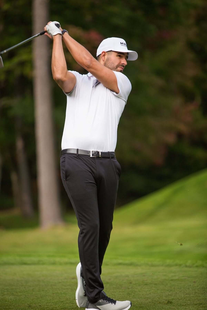Homme jouant au golf, portant un polo blanc et un pantalon noir sur un terrain verdoyant.