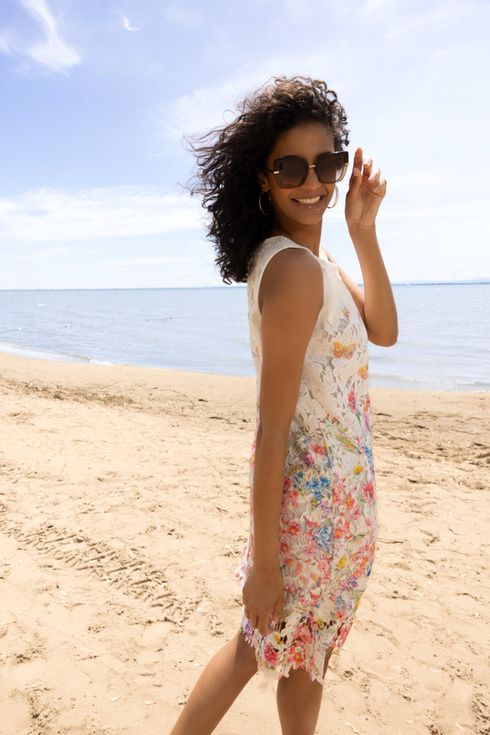 Femme sur une plage portant une robe florale avec détails, dentelle délicate et allure estivale élégante, idéale pour occasions spéciales.