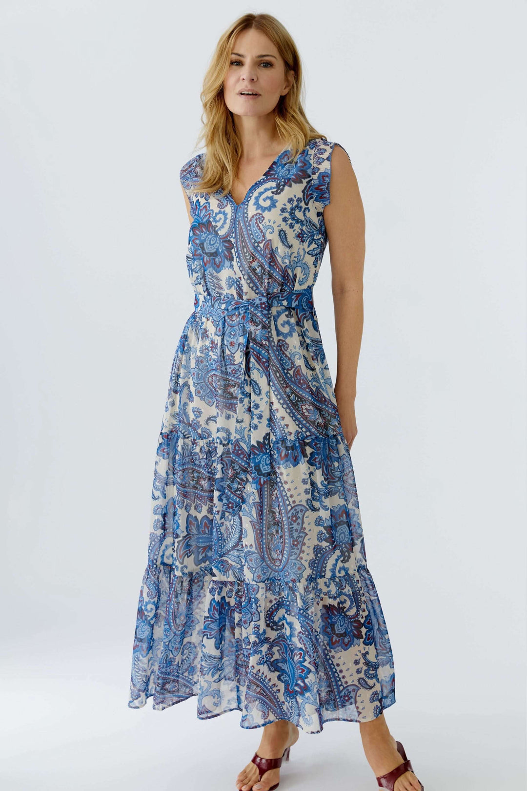 Femme portant une robe longue à imprimé paisley bleue, sans manches, encolure en V, jupe à volants étagés, ceinture à nouer.