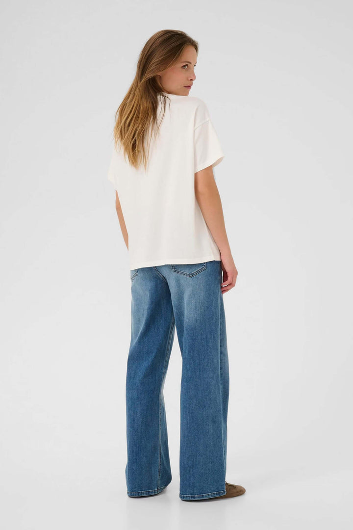 Femme portant un t-shirt Helen blanc avec un jean bleu, vue arrière, illustrant un style décontracté et essentiel pour la garde-robe.