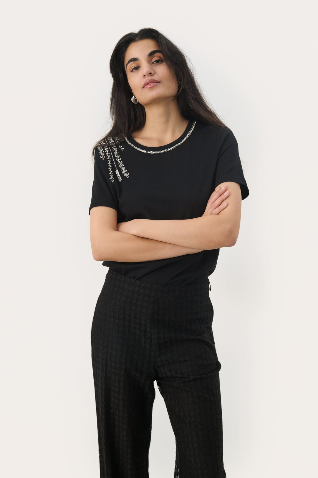 T-shirt noir à col rond avec broderies argentées et perles, associé à un pantalon noir, style élégant.