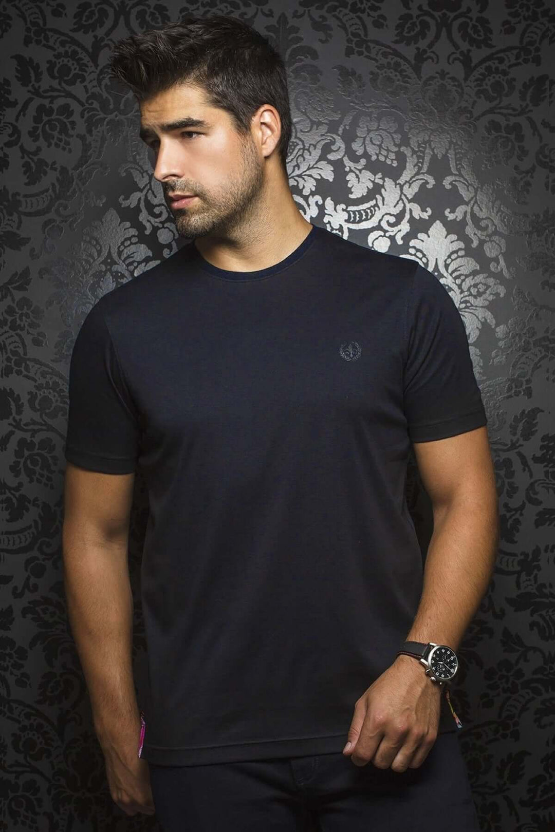 Homme portant un t-shirt à col rond noir, style minimaliste, avec logo brodé, sur fond mural stylisé.